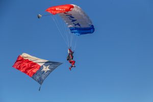 RE/MAX of Texas Parachute Team skydiving onto the launch field at the H-E-B | Central Market Plano Balloon Festival with the Texas flag.